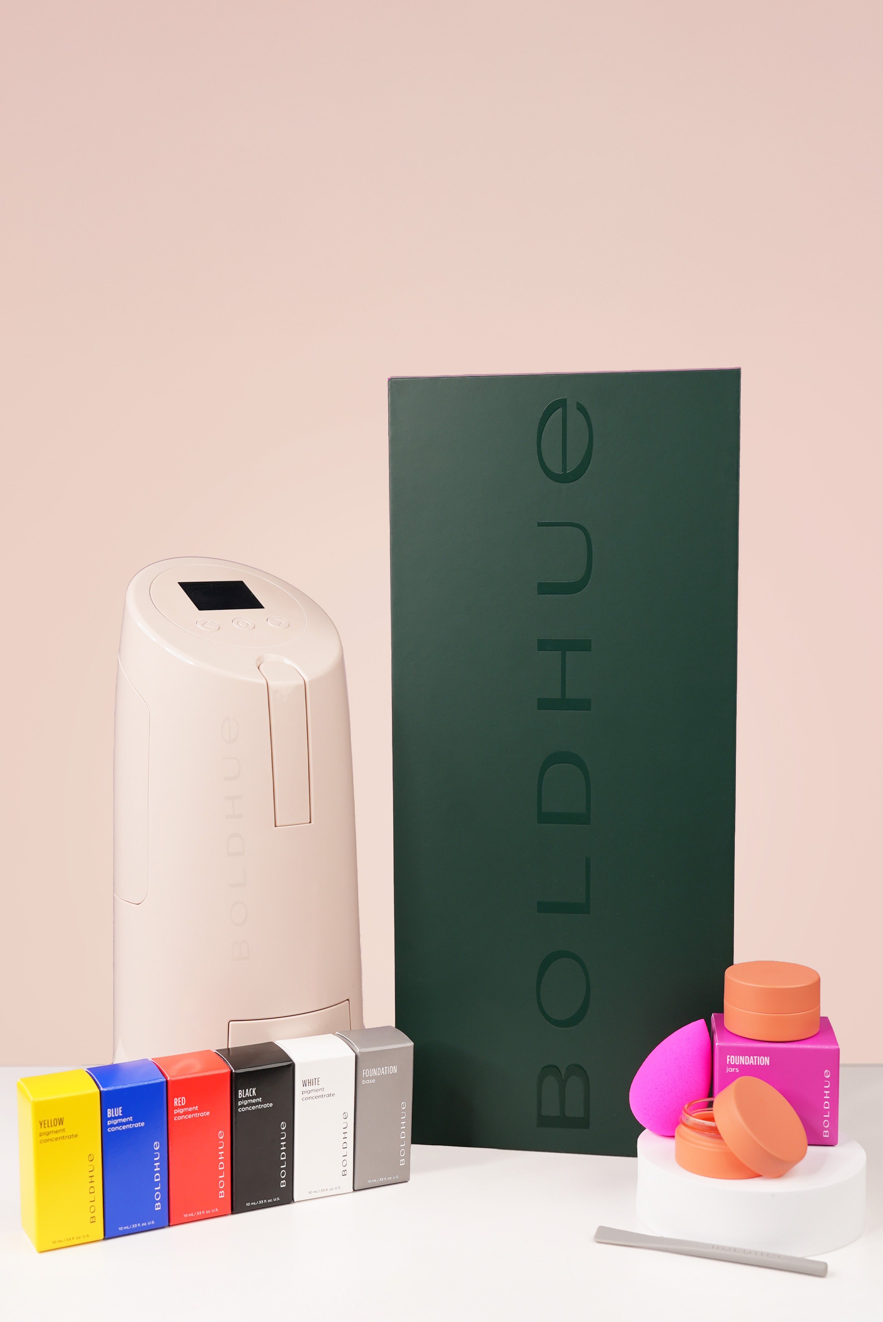 a picture of the boldhue starter kit that include the device, six pigment cartridges in base, black, white, red, yellow and blue needed to run the device, an accessory kit of two storage jars, a mixing spatula and a beautyblender to apply the foundation